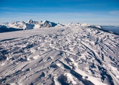 Vast snow-covered mountain slope with ski tracks and distant peaks under a blue sky.