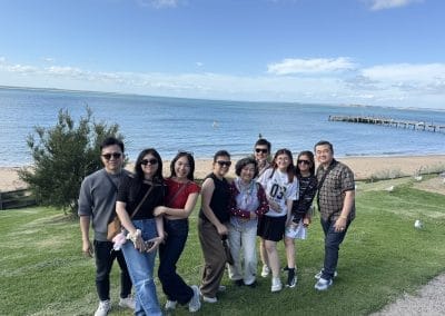 Group of people standing on grassy area near a sandy beach with a pier stretching into the water under a partly cloudy sky