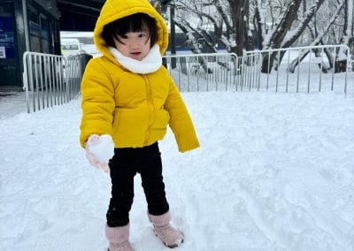 Child in yellow jacket holding a snowball in the snow.