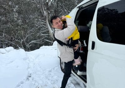 A man holding a child in a yellow jacket, both smiling, beside a white van in a snowy landscape with trees