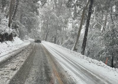 A snow-covered road winding through a forest, with a vehicle in the distance